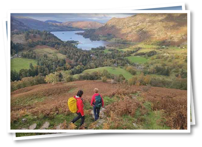 Two people walking down a fell side in the Lake District, surrounded by rolling hills and scenic countryside